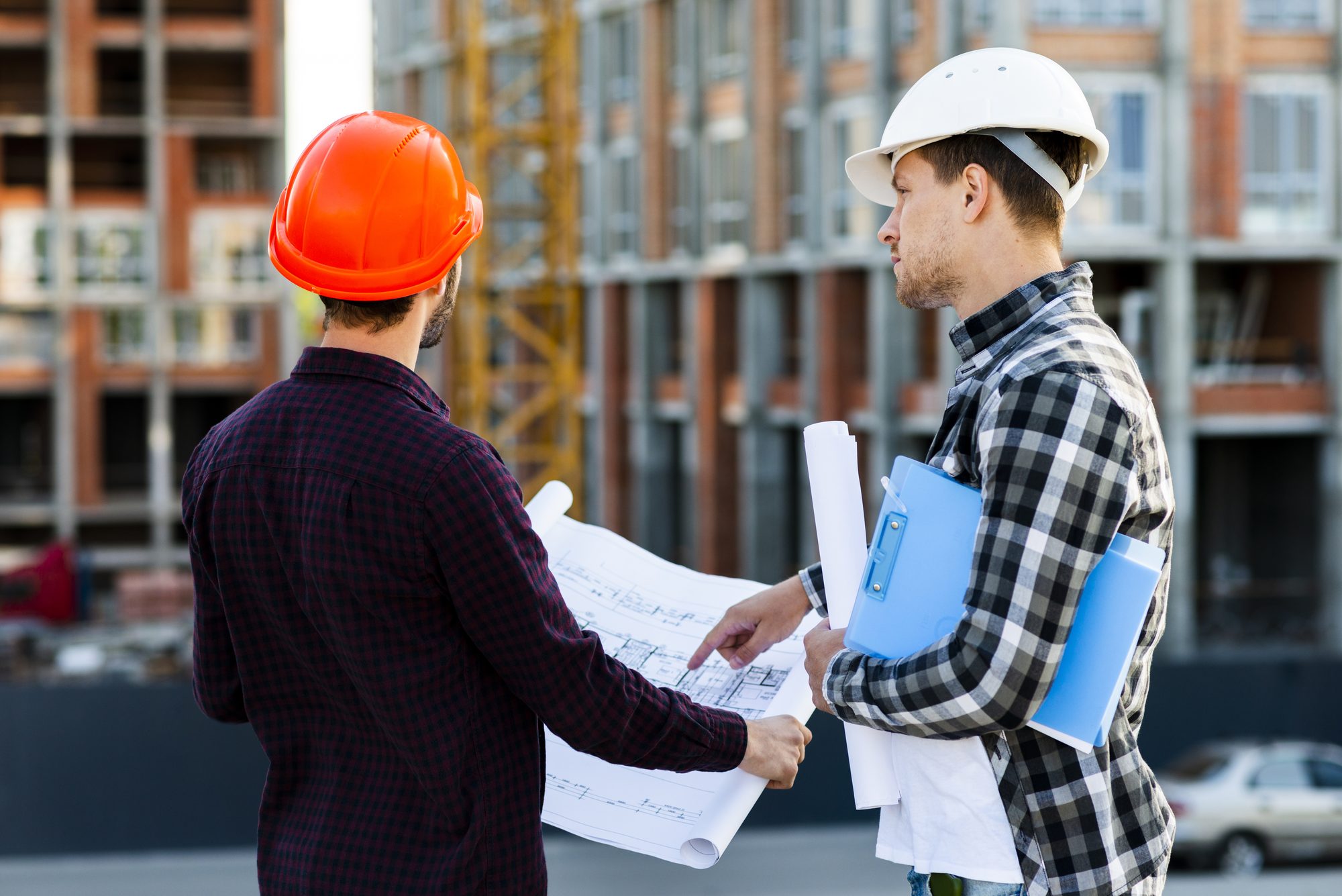 Two site engineers in hard hats reviewing drawings in front of an apartment block under construction
